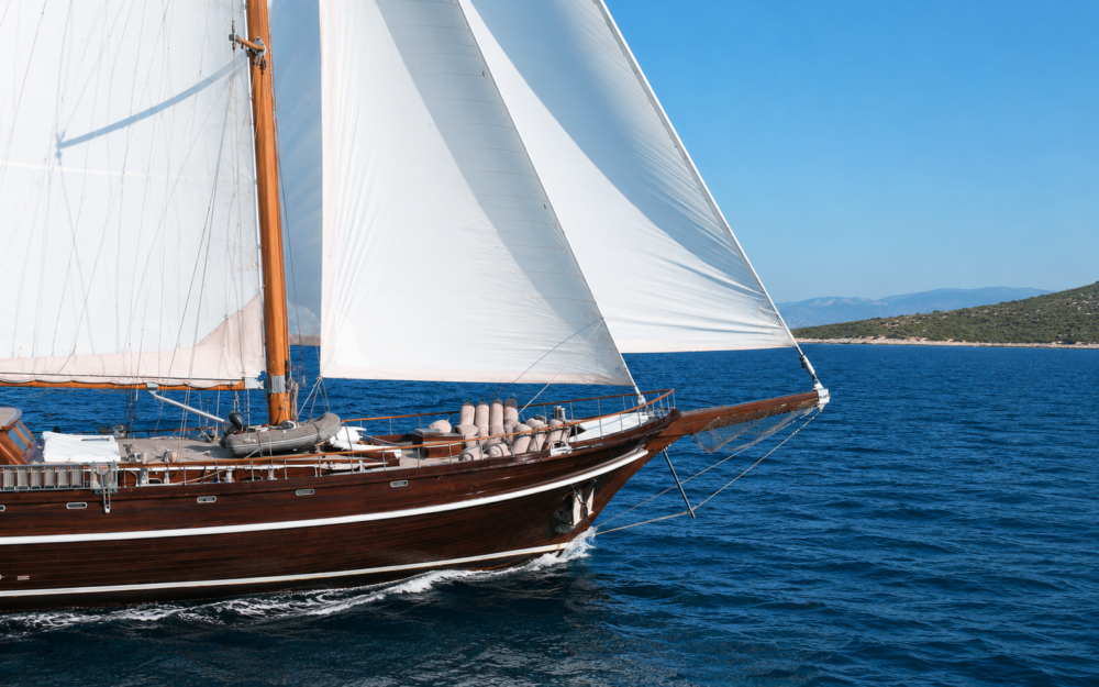 Close-up view of a luxury dark brown Turkish gulet sailing on calm deep blue waters under bright white sails during a sunny Mediterranean day. Elegant wooden yacht detail with clear skies, vibrant sea colors, and a refined classic design.