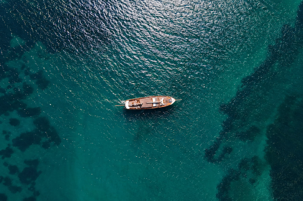 Top-down aerial view of a small wooden boat sailing alone over clear turquoise-green deep sea water, with sunlight reflecting on gentle waves and darker underwater patches